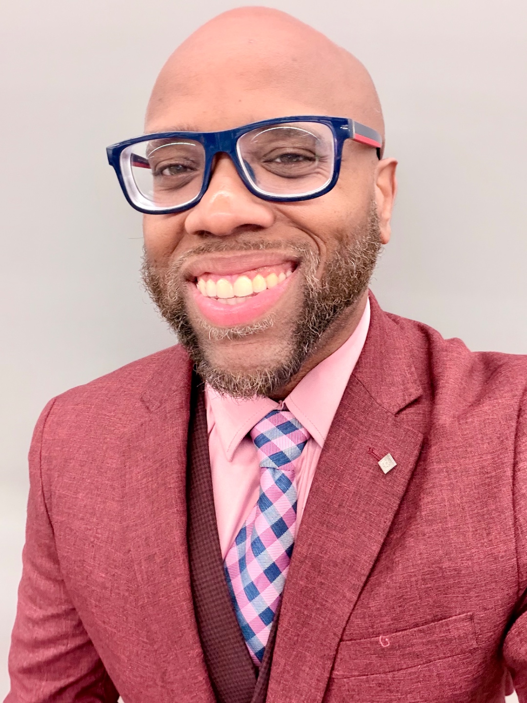 Miles Randall in a rose blazer, pink shirt, pink-and-navy check tie, and bold navy-and-red glasses, smiling in a studio portrait.