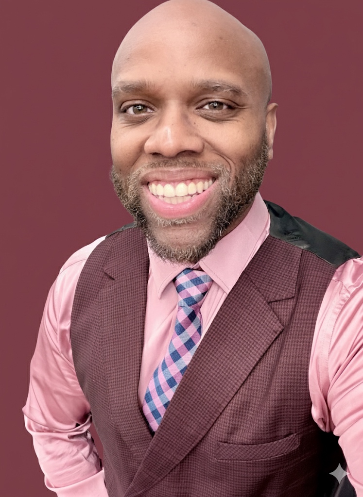 Miles Randall in a burgundy waistcoat, pink shirt, and pink-and-blue check tie against a rich burgundy studio backdrop.
